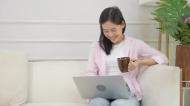 Young asian business woman work from home with laptop computer and drinking coffee on sofa in living room, freelance girl using notebook sitting on couch with comfort and relax, lifestyles concept.