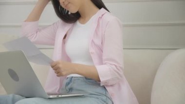 Young asian woman stress sitting on sofa reading notification document about loan and expense finance while worried and unhappy and shocked at home, financial and invoice letter, business concept.
