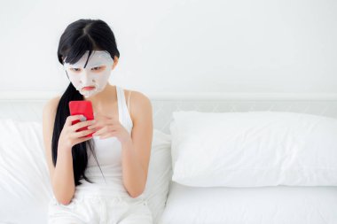 Yüzünde çarşaf maskesi olan güzel Asyalı bir kadın yatak odasında akıllı telefon izleyip cilt bakımı, cilt bakımı, sağlık ve sağlık için yüz maskesi takıyor..