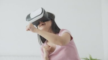 VR kulaklık takan genç Asyalı bir kadın evdeki yatak odasındaki yatakta heyecanla oyun oynuyor. Sanal gerçeklik ya da metaz yeniliklerini kullanarak 3 boyutlu, yaşam tarzları ve teknoloji kavramını kullanıyor..