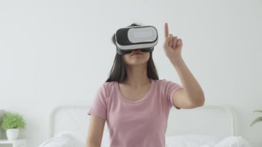VR kulaklık takan genç Asyalı bir kadın evdeki yatak odasındaki yatakta heyecanla oyun oynuyor. Sanal gerçeklik ya da metaz yeniliklerini kullanarak 3 boyutlu, yaşam tarzları ve teknoloji kavramını kullanıyor..