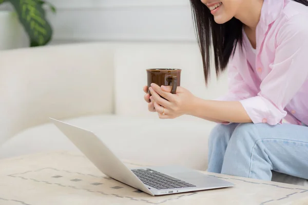 Young asian business woman work from home with laptop computer and drinking coffee on sofa in living room, freelance girl using notebook sitting on couch with comfort and relax, lifestyles concept.