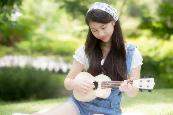 快乐的年轻亚洲女人在公园里坐着玩四弦琴 而休闲娱乐则是夏天的业余爱好 而女孩则是音乐家 带着积极的感觉和生活方式的观念做着活动 — 图库照片