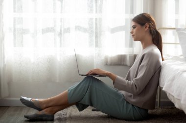 Güzel genç bir kadın yatağında dizüstü bilgisayarını kullanarak evdeki yatak odasında rahat ve rahat bir şekilde oturuyor. Defter üzerinde çalışan serbest bir kadın, bir kişi, yaşam tarzı konsepti..