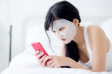 Yüzünde çarşaf maskesi olan güzel Asyalı bir kadın yatak odasında akıllı telefon seyrediyor. Güzellik kızı cilt bakımı, cilt bakımı, sağlık ve sağlık için yüz maskesi takıyor..