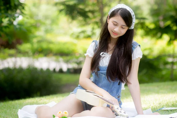 快乐的年轻亚洲女人在公园里坐着玩四弦琴 而休闲娱乐则是夏天的业余爱好 而女孩则是音乐家 带着积极的感觉和生活方式的观念做着活动 — 图库照片