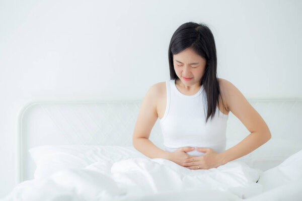 Young asian woman sitting on bed pain stomach or bowel with disease constipation in the bedroom at home, unhappy female sick and menstruation and stomachache, medical and health concept.
