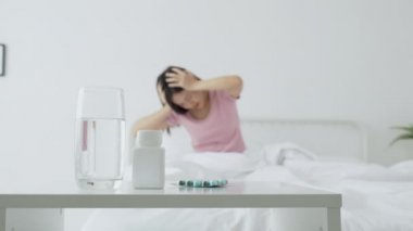 Genç Asyalı kadın yatak odasında yatak odasında ilaç ve su içerken baş ağrısı, mutsuz kadın yorgunluğu ve hasta baş ağrısı, ilaç tedavisi, tıbbi ve sağlık konsepti....