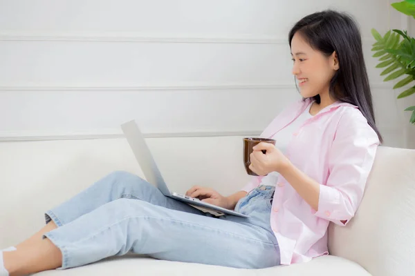 Young asian business woman work from home with laptop computer and drinking coffee on sofa in living room, freelance girl using notebook sitting on couch with comfort and relax, lifestyles concept.