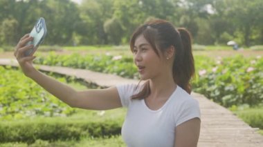 Beautiful young asian woman using smart phone taking a selfie while relaxation in the park, female using smartphone take a shooting picture in the garden, lifestyles and communication concept.