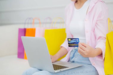 Young asian woman sitting on sofa using laptop computer shopping online with credit card while paper bag on sofa, female paying with transaction financial, purchase and payment, business concept.