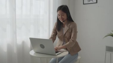 Young asian businesswoman wearing headset video call and explain conference online at home, woman wearing headphone meeting and talking for working from home, business and communication concept.