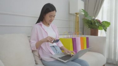 Kanepede oturan Asyalı genç bir kadın internet üzerinden dizüstü bilgisayar alışverişi yapıyor. İnternete kredi kartı alıyor, alışveriş, alım-satım ve ödeme ile ödeme yapan mutlu bir kız..