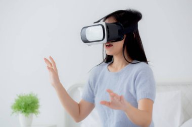 VR kulaklık takan genç Asyalı bir kadın evdeki yatak odasındaki yatakta heyecanla oyun oynuyor. Sanal gerçeklik ya da metaz yeniliklerini kullanarak 3 boyutlu, yaşam tarzları ve teknoloji kavramını kullanıyor..