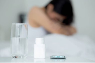 Young asian woman is depression and having mental health problem sitting on bed while drug and water on desk in the bedroom, emotion and negative, woman and expression, medical and health concept.