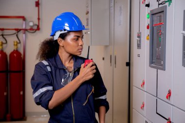 Electrical young asian woman engineer examining maintenance cabinet system electric and using radio in control room at industrial factory, technician or electrician inspection power distribution.