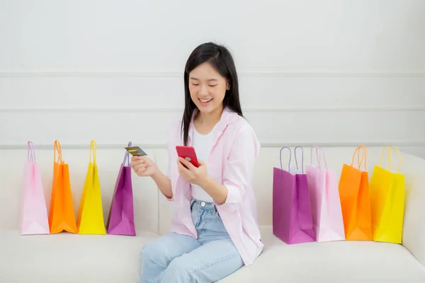 Young asian woman sitting on sofa using smart phone shopping online with credit card while paper bag on couch, happy female paying with transaction financial, purchase and payment, business concept.
