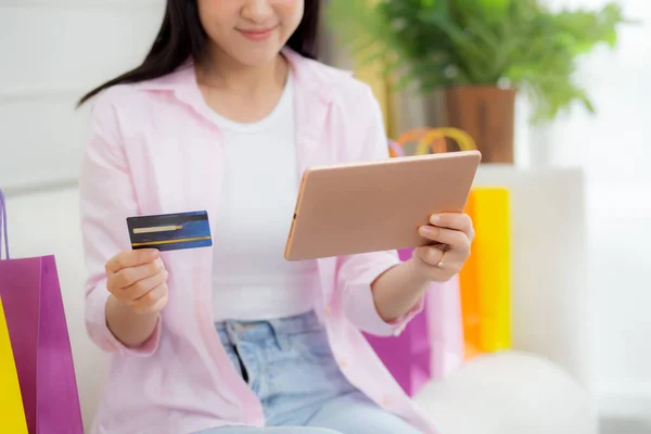Young asian woman sitting on sofa using digital tablet shopping online with credit card while paper bag on couch, female paying with transaction financial, purchase and payment, business concept.