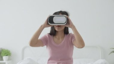 VR kulaklık takan genç Asyalı bir kadın evdeki yatak odasındaki yatakta heyecanla oyun oynuyor. Sanal gerçeklik ya da metaz yeniliklerini kullanarak 3 boyutlu, yaşam tarzları ve teknoloji kavramını kullanıyor..