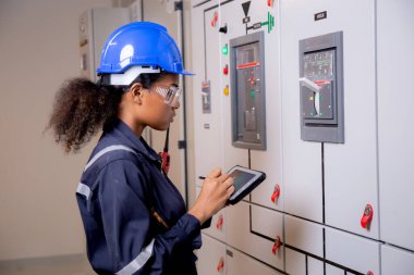 Electrical young asian woman engineer examining maintenance cabinet system electric and using tablet in control room at industrial factory, technician or electrician inspection power distribution.