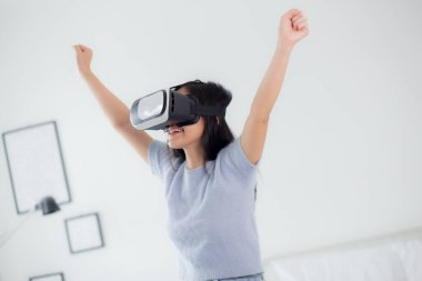 Young asian woman wearing vr headset playing game with excited in the bedroom at home, female using virtual reality or metaverse innovation for simulation 3D, lifestyles and technology concept.
