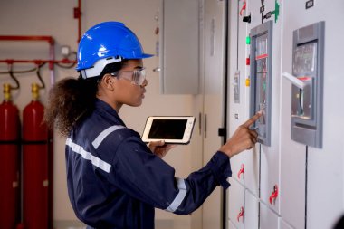 Electrical young asian woman engineer examining maintenance cabinet system electric and using tablet in control room at industrial factory, technician or electrician inspection power distribution.
