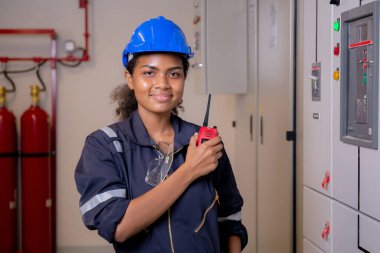 Electrical young asian woman engineer examining maintenance cabinet system electric and using radio in control room at industrial factory, technician or electrician inspection power distribution.