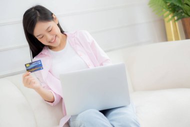 Young asian woman sitting on sofa using laptop computer online shopping with credit card, happy female payment with debit card on couch, purchase and payment, business and technology concept.