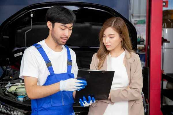 Young mechanic talking to customer asian woman about car with repairing in garage, auto service, technician man explaining problem of vehicle and look clipboard with client, transportation concept.