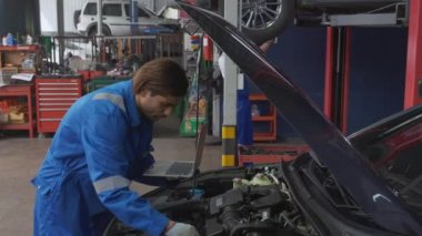 Young man is mechanic using laptop computer checking engine of car in the garage, worker examining and diagnostic automobile, technician and maintenance of vehicle, transport concept.