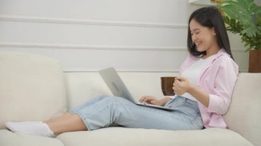 Young asian business woman work from home with laptop computer and drinking coffee on sofa in living room, freelance girl using notebook sitting on couch with comfort and relax, lifestyles concept.
