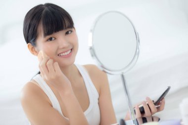 Güzel, genç Asyalı bir kadın yanak makyajına pudra üflüyor kozmetik aynası, cilt yüzlü ve yatak odasındaki kırışıklıklar, kadın kozmetolojisi, cilt konsepti..