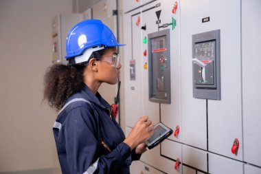 Electrical young asian woman engineer examining maintenance cabinet system electric and using tablet in control room at industrial factory, technician or electrician inspection power distribution.