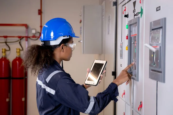 Electrical young asian woman engineer examining maintenance cabinet system electric and using tablet in control room at industrial factory, technician or electrician inspection power distribution.