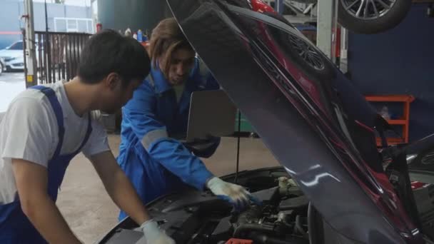2人の若い男は ガレージ内の車のラップトップコンピュータチェックエンジンを使用して整備士です 自動車を検査し 診断労働者 技術者と車両のメンテナンス 輸送コンセプト — ストック動画