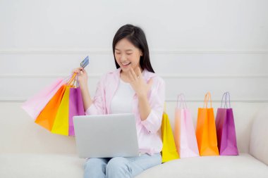 Kanepede oturan Asyalı genç bir kadın bilgisayarla online alışveriş yapıyor. Kredi kartıyla ve kesekağıdıyla ödeme yapıyor. İşletme konseptiyle ödeme yapan mutlu bir kadın..
