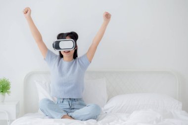 Young asian woman wearing vr headset playing game with excited in the bedroom at home, female using virtual reality or metaverse innovation for simulation 3D, lifestyles and technology concept.
