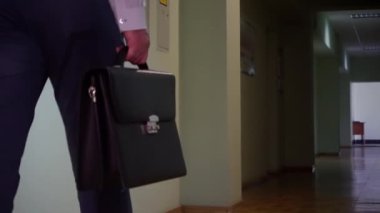 Pantolon ve gömlek giymiş bir işadamı elinde siyah bir çantayla koridorda yürüyor. Ofiste yürüyen bir adamın elindeki evrak çantasının yakın çekimi..
