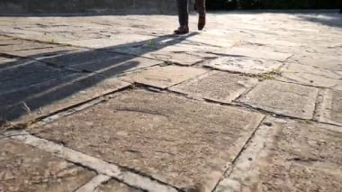 Taş yolda yürüyen bir adamın gölgesi. Ayakkabılı ve pantolonlu bir adamın bacaklarının yakın çekimi..