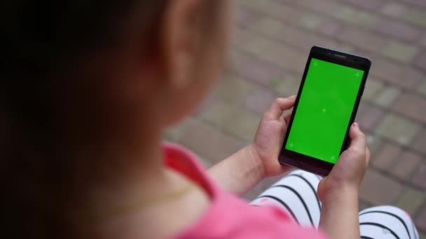 夏の夜公園のベンチに座って手に緑の画面のスマートフォンを持っている子供の女の子 スマートフォンは クロマキーディスプレイとコンセプトをモックアップ — ストック動画