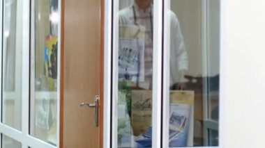 Genç bir adam cam şeffaf duvarlarla koridorda yürür ve kapıyı açar. Kapı kolunu çeviren bir ofis çalışanının yakın çekimi. Camdaki bir adamın yansıması..