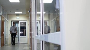 Genç bir ofis çalışanı koridor boyunca yürür kapıyı açar ve içeri girer. Ofis kapısını açan adamın camında yansıma var..