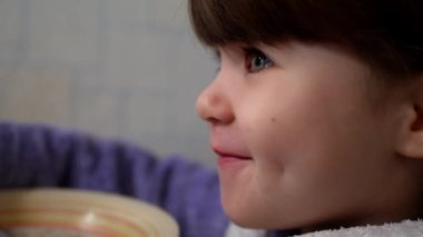 Tatlı küçük bir kız kahvaltıda dikkatlice sıcak süt lapası yiyor. Çocuk yemekten önce sıcak bir kahvaltı üfledi. Sağlıklı kahvaltı ve ev hayatı.