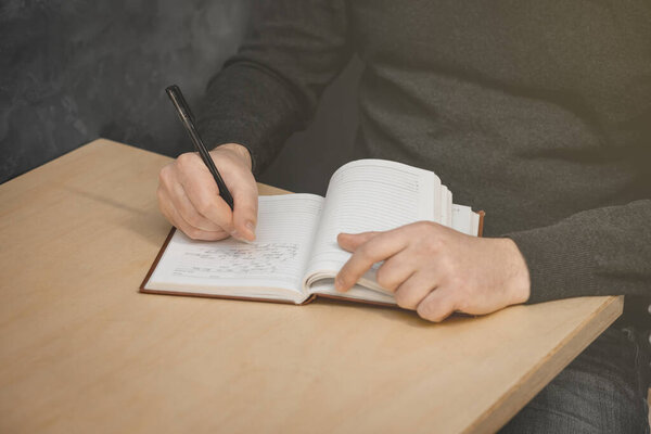 The man's hand holds a pen and writes in a notebook. The second hand is holding the notebook.
