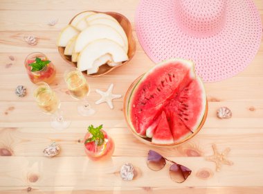Kavun dilimleri, karpuz, pembe şapka, güneş gözlükleri, şampanya bardakları, deniz balıkları, deniz kabukları ve ahşap masada serinletici yaz karpuzu limonatası. Üst görünüm.