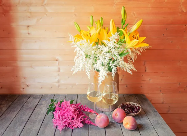 泥土瓶中的黄色百合花和白色的子午线花束 白葡萄酒杯 粉红色的子午线和陈年老旧木桌上的新鲜水果 图库照片