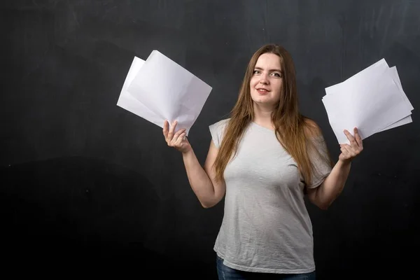 A woman teacher in light clothes stands with sheets of paper in her hands against a black wall. Looking into the camera, vertical photo. place for text