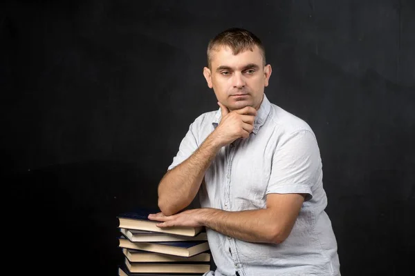 a man of European appearance is standing near the books with a thoughtful look. look at the floor. teacher on the background of a black slate wall.