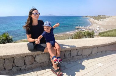 Koyu saçlı, beyaz bir kadın, tatilde kucağında bebekle bir tatil köyünde güzel masmavi deniz suyunun ve dağların önünde oturuyor.