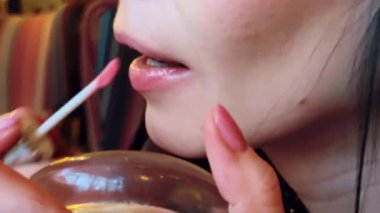 Koyu saçlı, beyaz bir kızın pembe parlatıcıyla dudak makyajı yaptığı yakın plan.